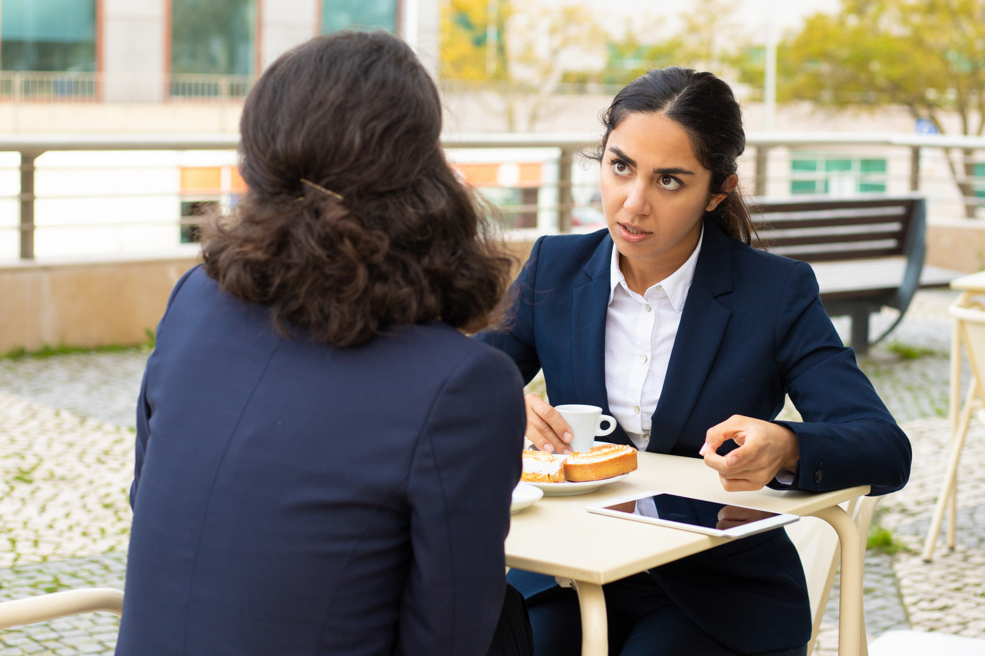 two business leaders in suits having an intent coaching style conversation at an outdoor café with coffee pastries and a tablet on the table   dr krishna athal